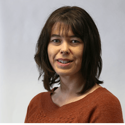 Woman with dark hair and bangs, smiling, wearing a brown sweater against a white background.